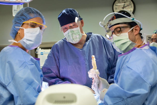 Three doctors look towards a machine in the foreground while performing surgery. All three are in scrubs, masks, and glasses.