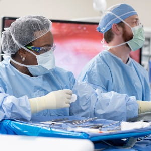 A woman in scrubs, mask, and glasses stands over a patient on the operating table with surgical instruments in front of her. A man in scrubs, mask, and glasses is behind her looking off to his left.