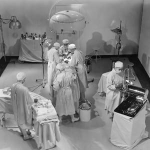 A view of an operating room from above. A patient is on the table surrounded by four people in scrubs while two other people tend to other tasks.
