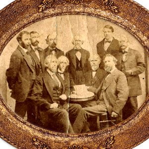 Ten men look at the camera, surrounding a table. The photo is wrinkled and in a circular frame.