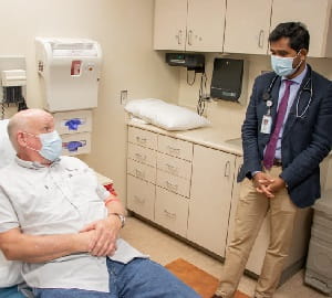 A man in a mask lies on a bed in a doctor’s office. The doctor, in a suit on the right, also wears a mask as the two are in a discussion.