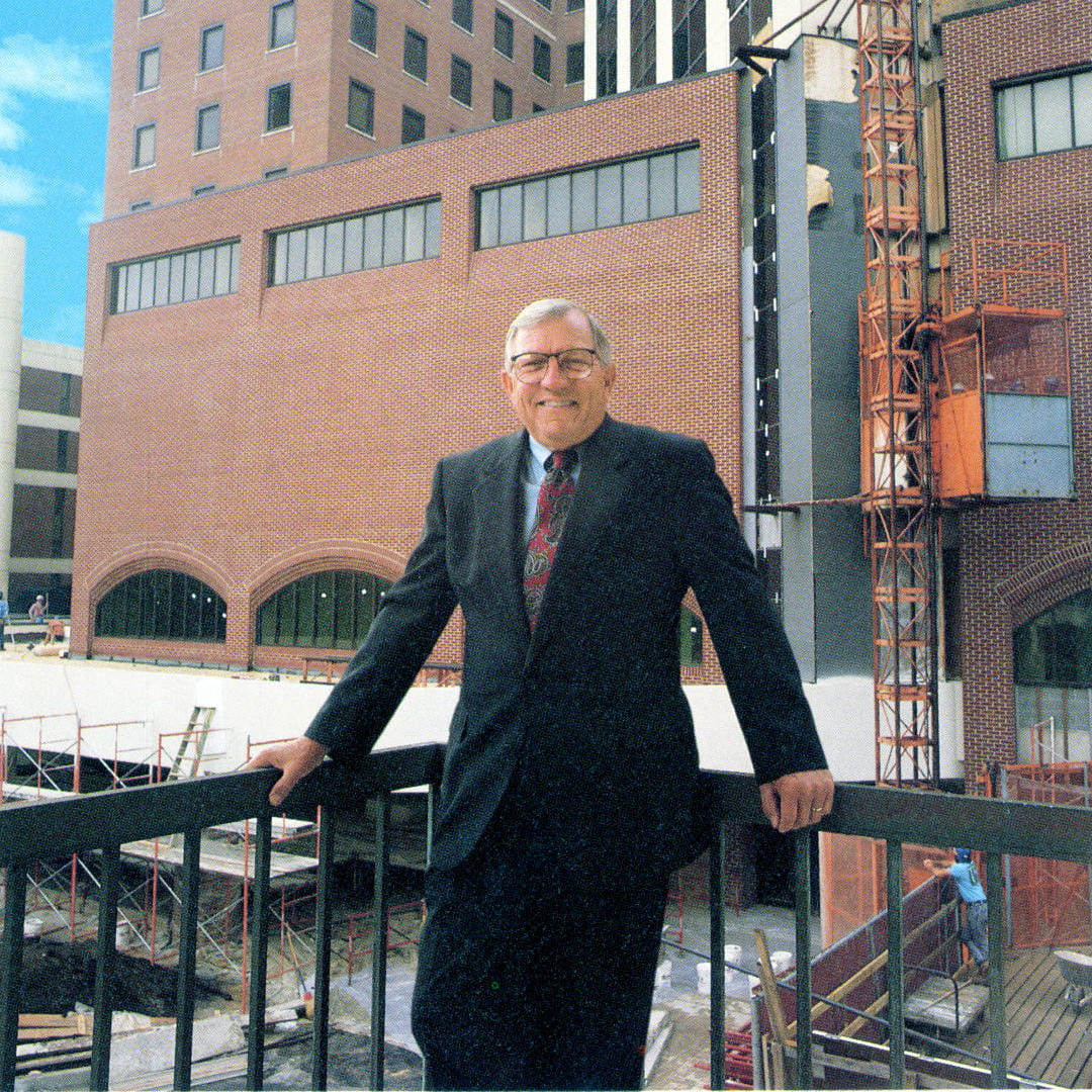 A man in a suit stands against a railing in front of a large brick building.