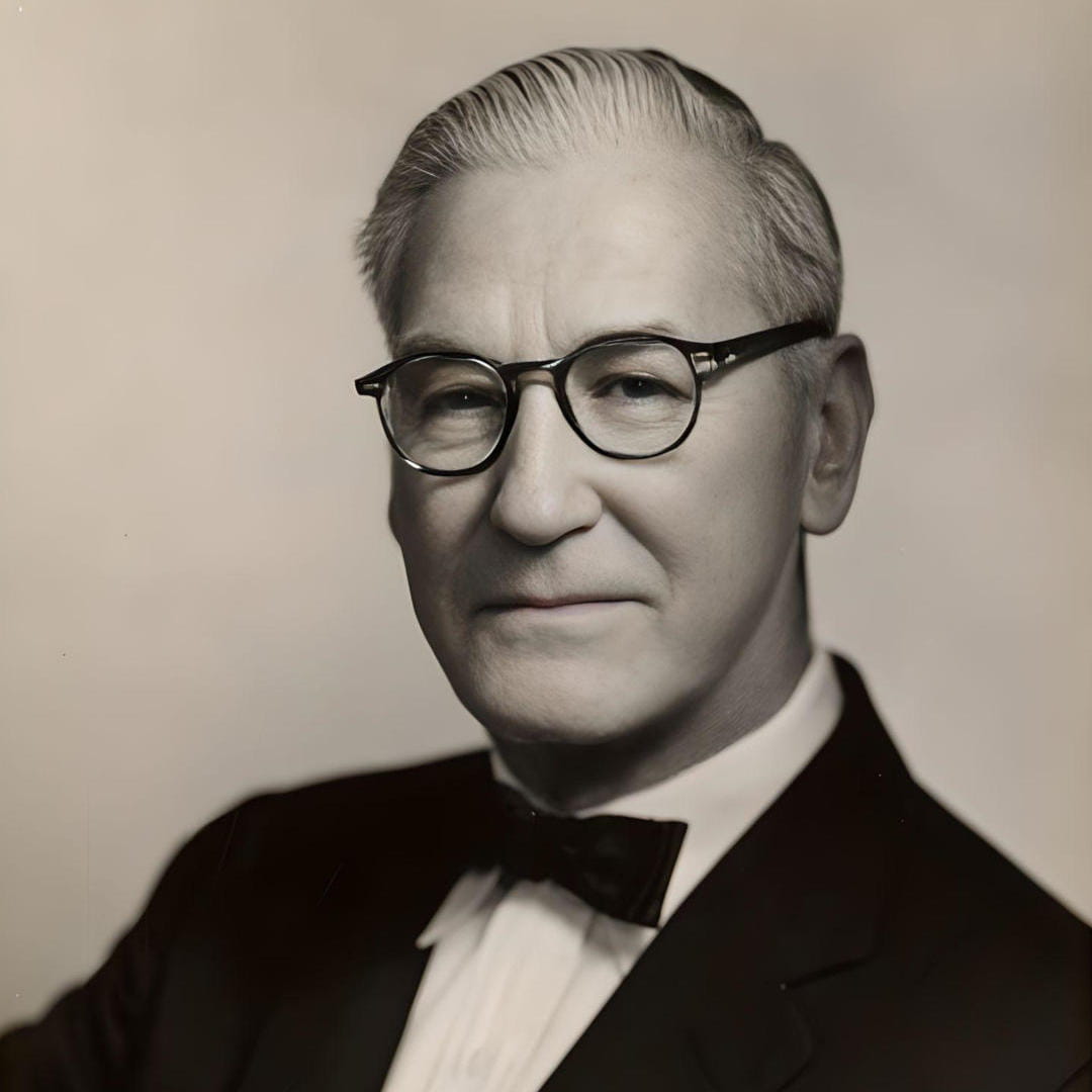 A man with slicked back hair and glasses looks toward us. He is wearing a dark coat, a white shirt, and a dark bowtie.