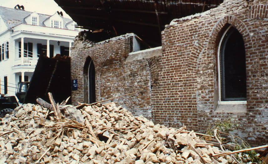 A building has had its roof torn off by a storm. In the foreground, a pile of bricks lay in front of a wall that remains.